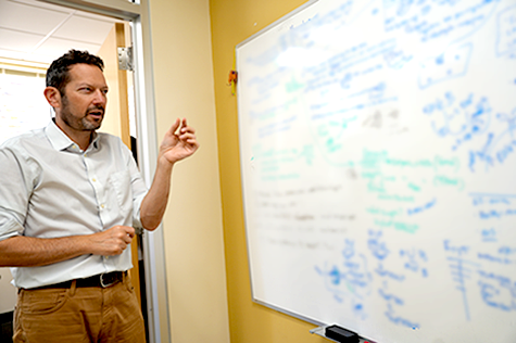 Image: Micah Luftig, Ph.D., standing beside a whiteboard covered with scientific notes and diagrams outside a laboratory office.