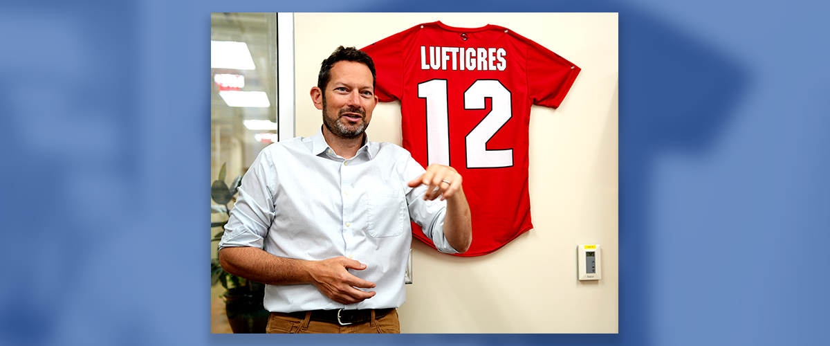 Image: Micah Luftig, Ph.D., standing in his office beside a red jersey mounted on a wall