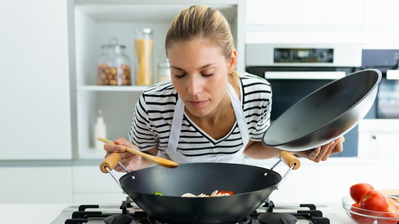 woman leaning over a wok, with the lid in one hand and a spoon in the other, sniffing what she's cooking.