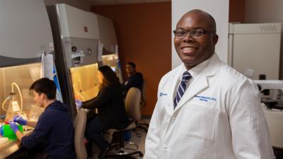 Dr. Olabisi in his lab, people working under safety hoods behind him. 