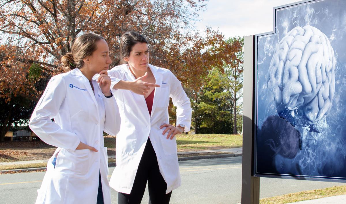 Jessica Waibl Polania (left) and Selena Lorrey, outside in their lab coats looking at a sign with the image of a brain on it. 