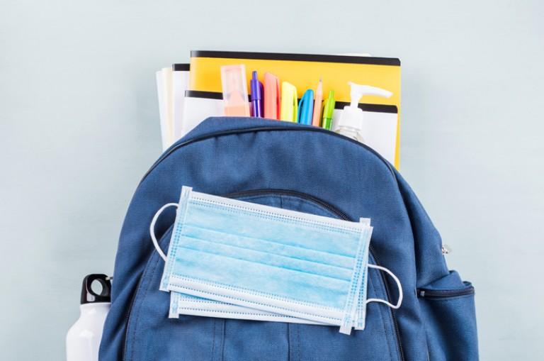 backpack with school supplies hanging out of it and a blue paper mask on top. 