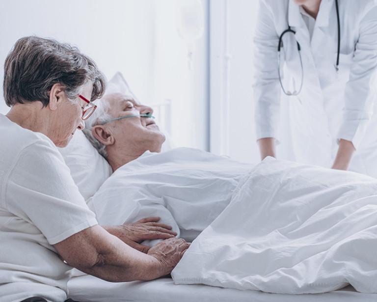 Elderly woman at the bedside of an elderly man in a hospital bed being treated by a healthcare worker.