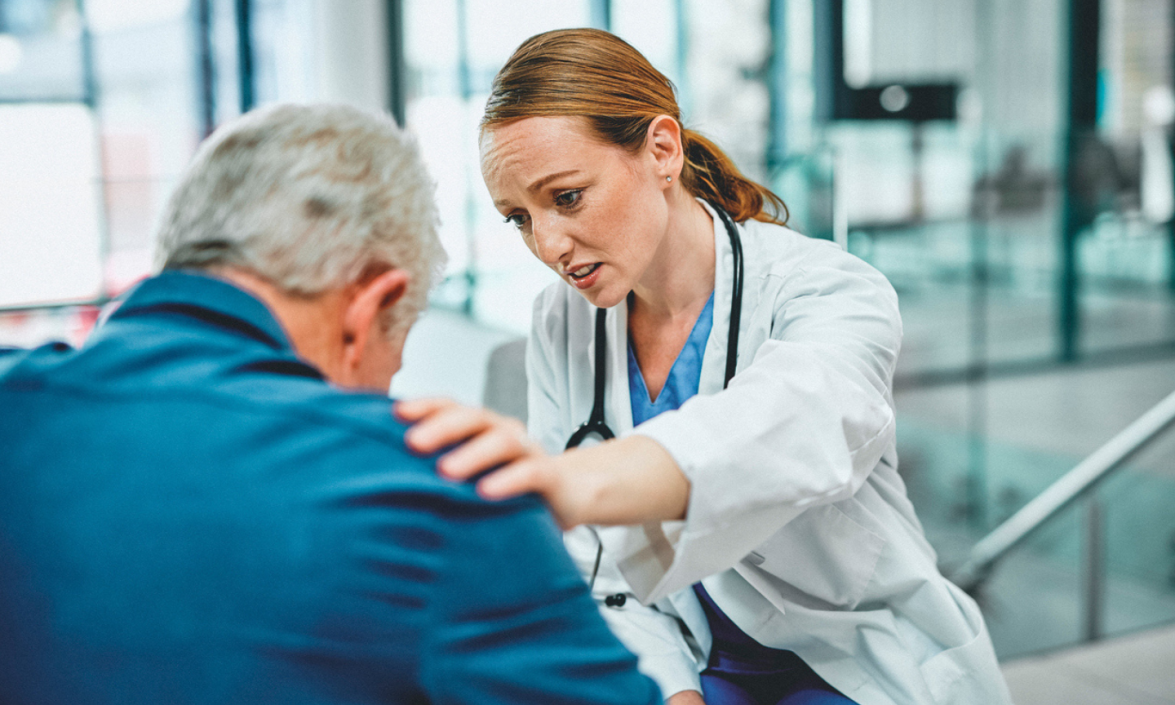 A woman in a white coat comforts an older man, putting a hand on his shoulder. 