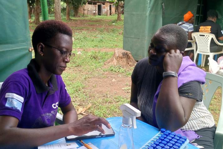 Two women sit at a table outdoors facing each other, one using a mobile phone.