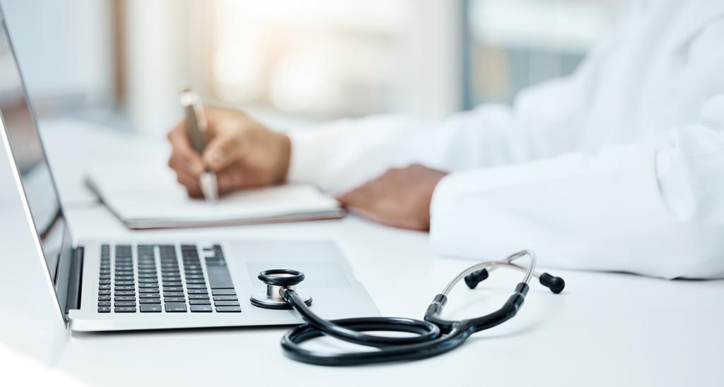 A person in a white coat sits at a laptop with a stethoscope nearby