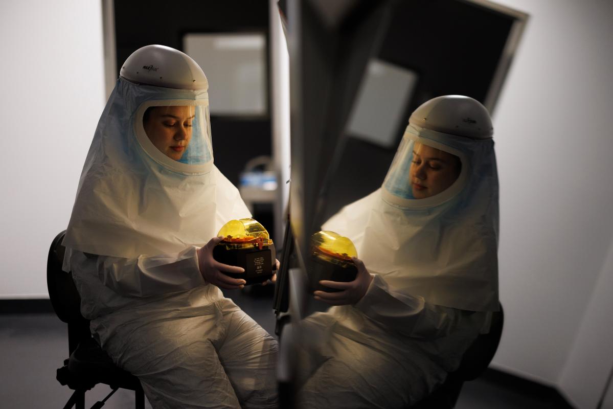 A researcher in full-coverage protective garb handles a sample beside a shiny surface that reflects her image.