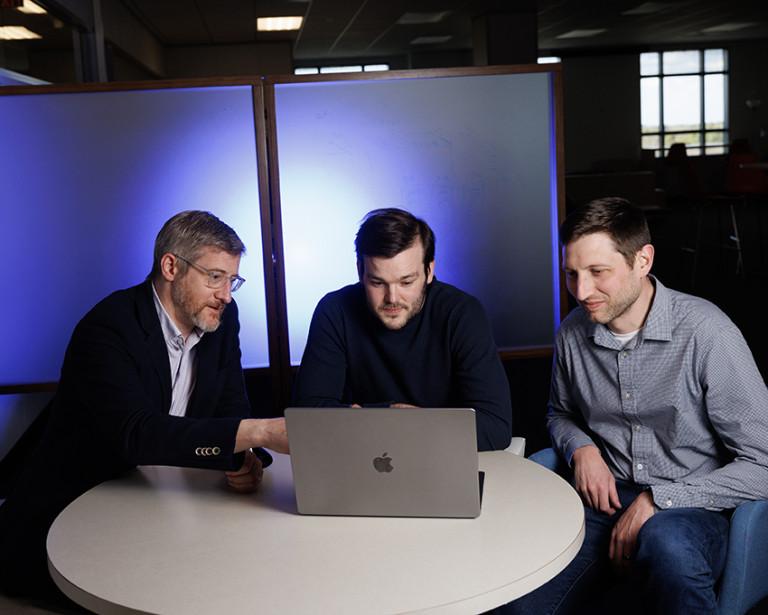 Duke researchers Jonathan Posner, Elliot Hill, and Matthew Engelhard sit at a small round table looking at a laptop screen