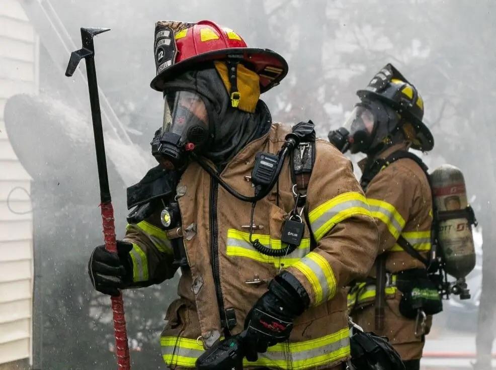 Two firefighters in heavy gear and gas masks fight a fire.