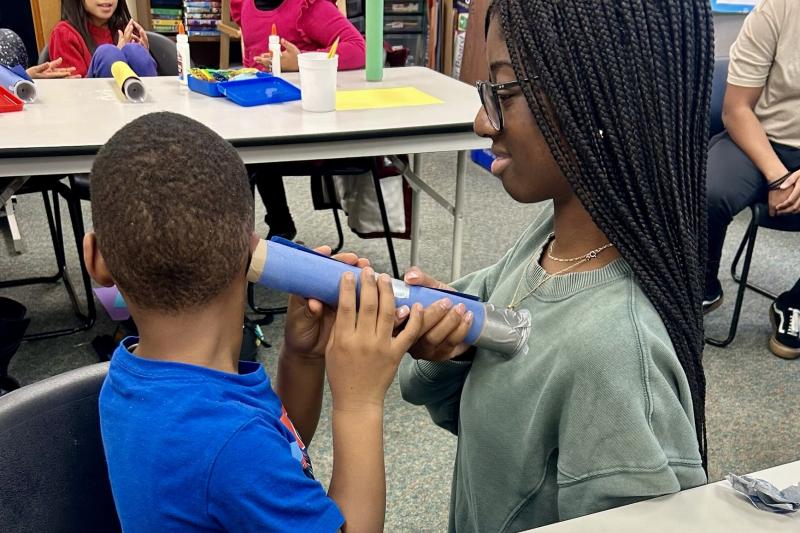 An elementary school student uses a homemade stethoscope to listen to a medical student's heart.