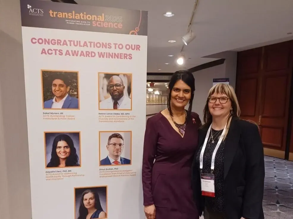 Gayathri Devi standing with ACTS Executive Director Kim Stelmaszak beside a large poster showing this year's ACTS Award winners