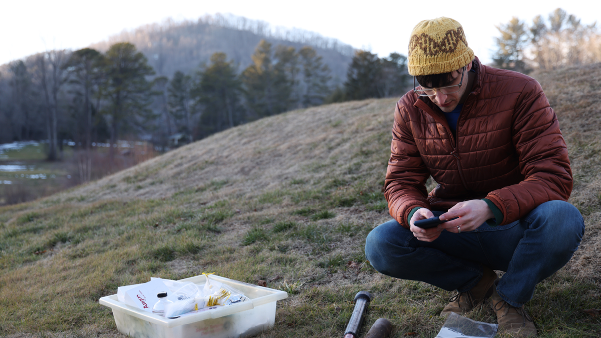 Male researcher in hat and puffy jacket, sitting on the side of a mountain collecting fungal samples. 