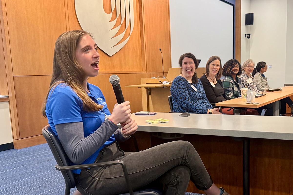 a student in the foreground speaking on a mic with faculty in the background laughing