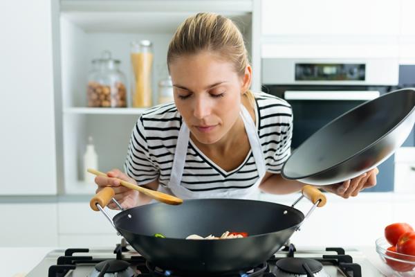 woman leaning over a wok, with the lid in one hand and a spoon in the other, sniffing what she's cooking.