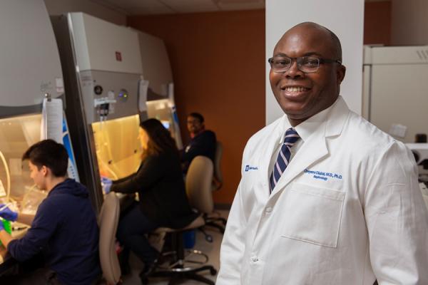 Dr. Olabisi in his lab, people working under safety hoods behind him. 