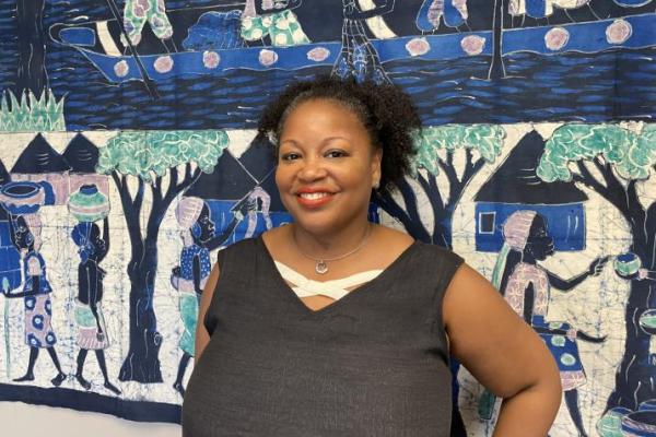 A Black woman stands in from of a blue, green, and black mural, smiling.