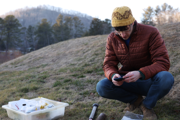 Male researcher in hat and puffy jacket, sitting on the side of a mountain collecting fungal samples. 