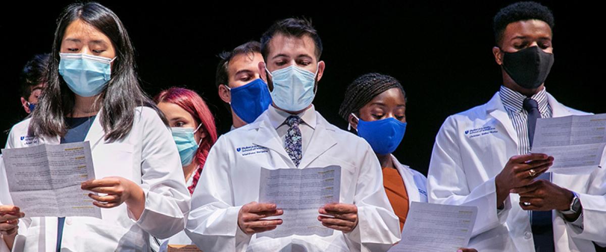 group of students in white coats read from papers at ceremony