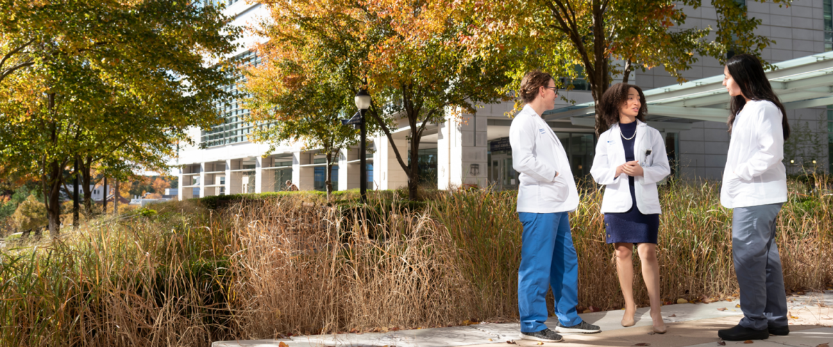 Three women med students in short white coats talking to each other while standing in front of the medical education building. 
