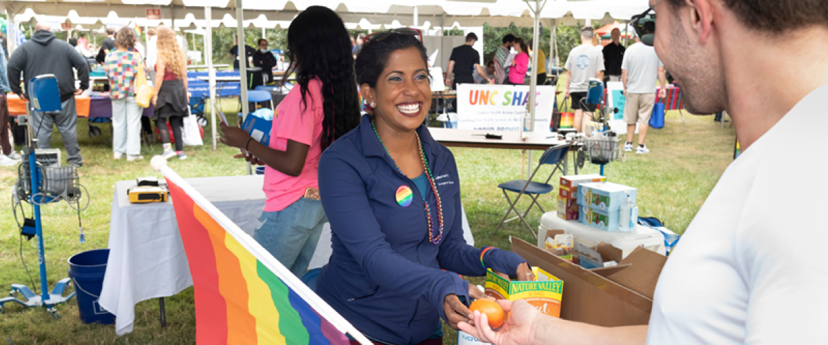 brown woman with a rainbow button handing out swag at a pride event.