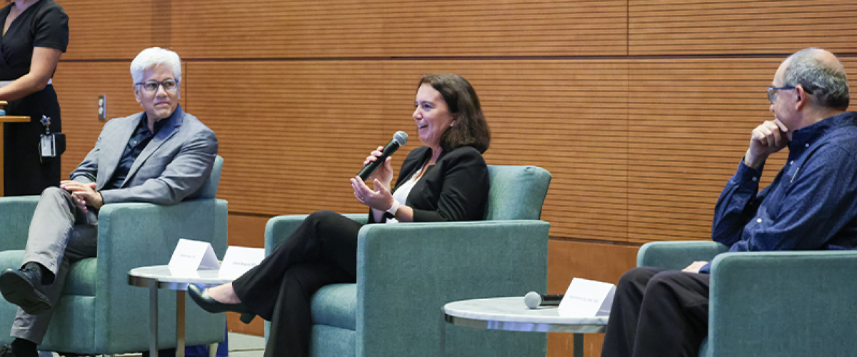 Three faculty sitting in chairs on a podium during a meeting