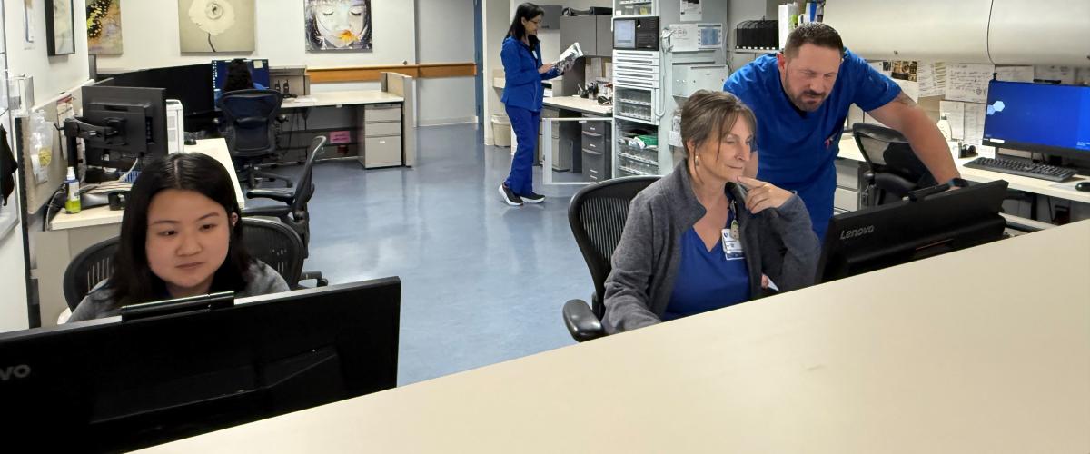 Clinical staff working in a hospital or research unit office space. Three staff members are seated at computer monitors at the front desk, with one staff member standing and providing assistance. In the background, another staff member in blue scrubs walks through the workspace. The room is well-lit with overhead fluorescent lights, and shelves and filing cabinets line the walls.