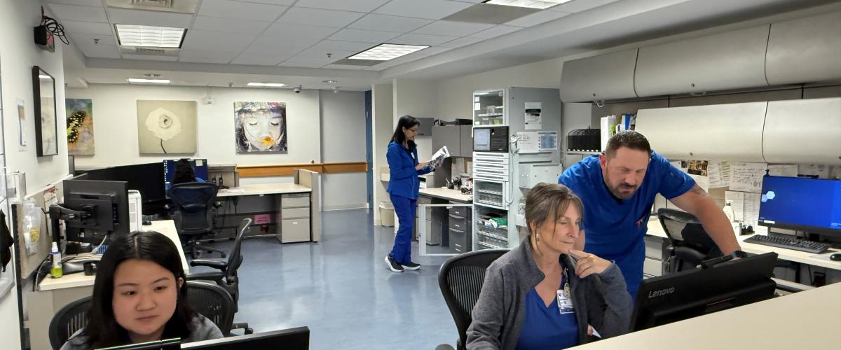 Clinical staff working in a hospital or research unit office space. Three staff members are seated at computer monitors at the front desk, with one staff member standing and providing assistance. In the background, another staff member in blue scrubs walks through the workspace. The room is well-lit with overhead fluorescent lights, and shelves and filing cabinets line the walls.