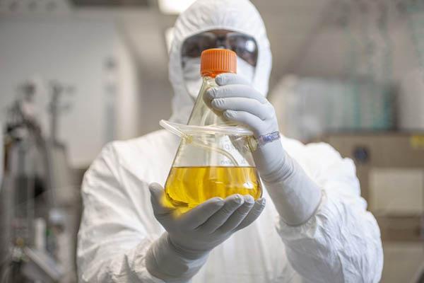 Researcher in total PPE (suite, gloves, mask & goggles) holds a capped flask up to the viewr