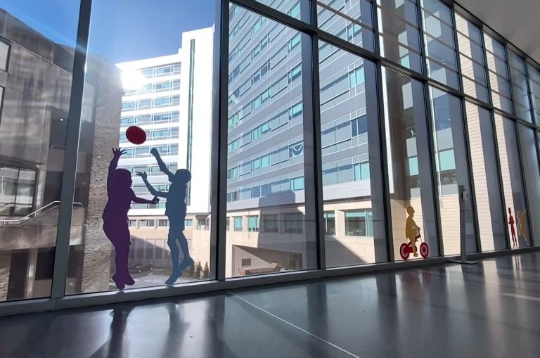 view out a window at the new hospital tower. Silhouettes of children playing with a ball affixed to the window in the foreground.