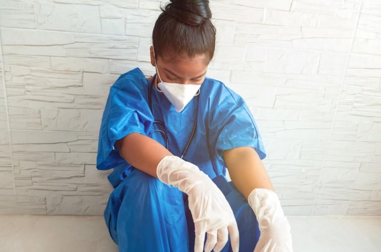black woman in scrubs and PPE slumped against a wall in exhaustion. 