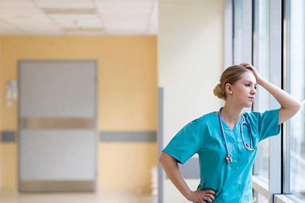 Woman in scrubs holding her head in exhaustion leaning against a window