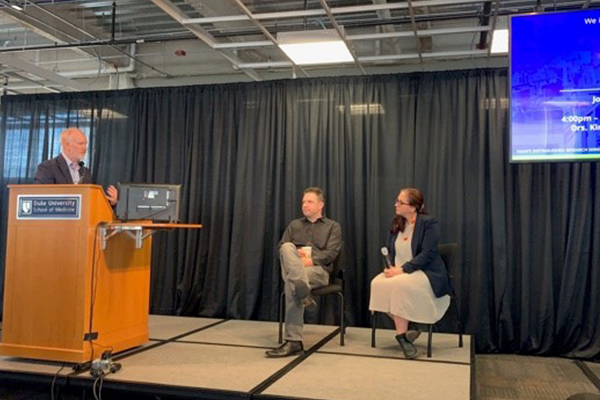 Colin Duckett behind a podium speaking with Cagla Eroglu, PhD and Andrew West, PhD