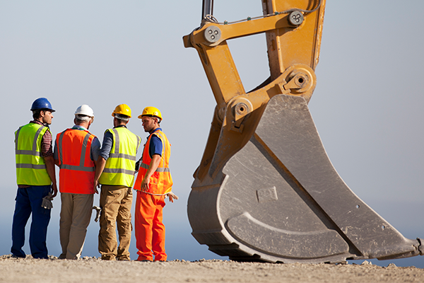 Four male construction workers in safety garb standing next to a giant backhoe shovel. 