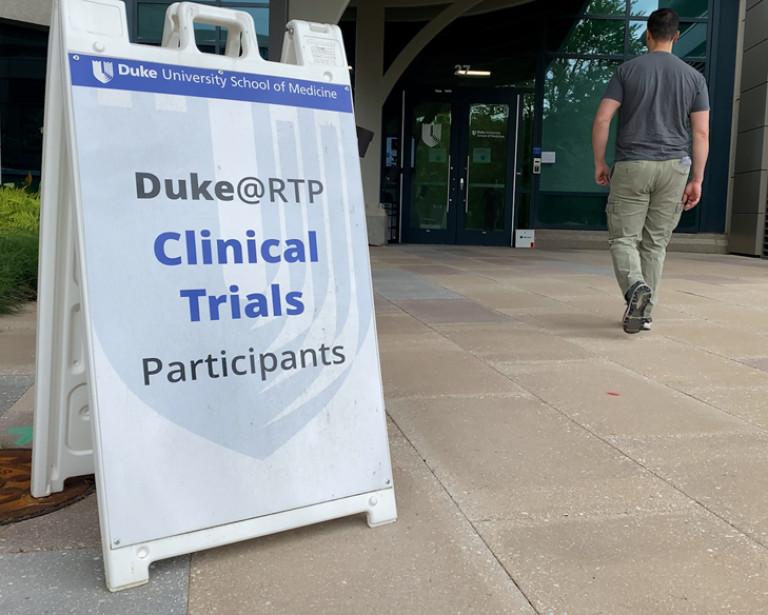 placard on a sidewalk "Duke@RTP Clinical Trials Participants. Man in the background entering a building.