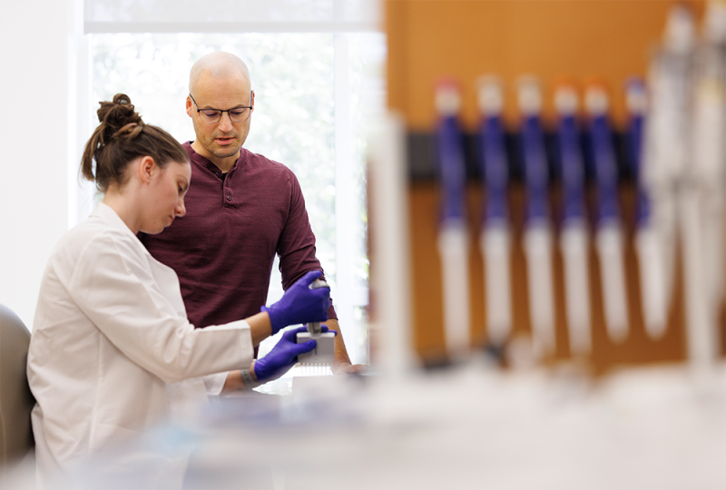 Jonathan Campbell with a gloved lab member watching her perform an experiment.