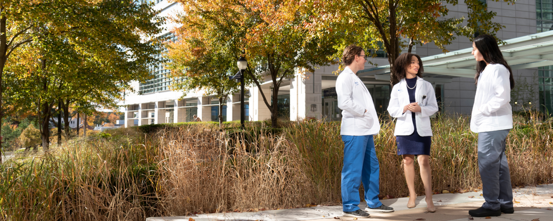 Three women med students in short white coats talking to each other while standing in front of the medical education building.