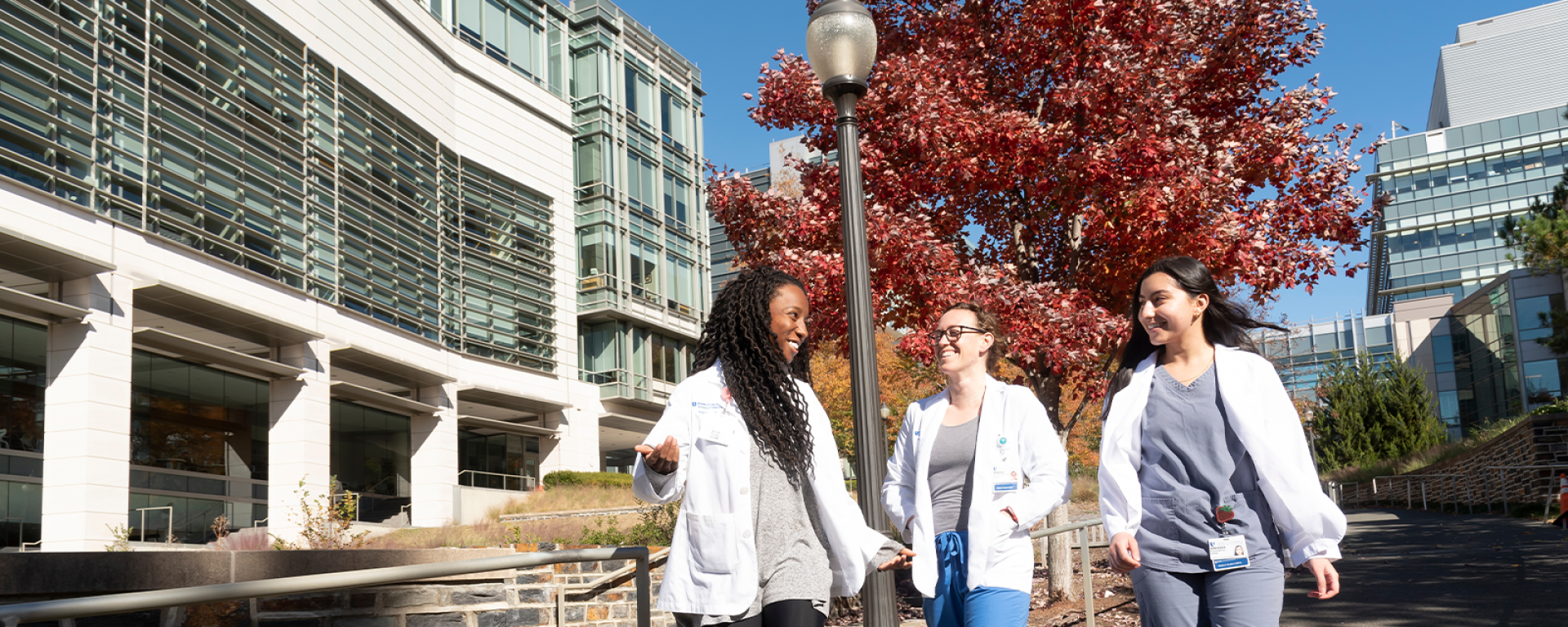 Three medical students walking next to the medical education building with colorful fall foliage in the background