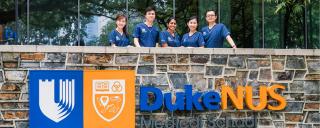 5 Medical students dressed in blue scrubs standing behind a waist high wall of Duke stone with the Duke-NUS logo on it. 