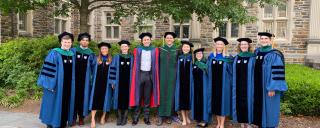 2022 MSTP Graduates in graduation robes with Director Chris Kontos in front of brick building and trees
