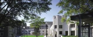 A photograph of a Duke Clinic courtyard surrounded by modern stone buildings. The scene includes large trees, green lawns, paved walkways, lampposts, and a few people walking. The bright blue sky and sunlight give the space a peaceful and welcoming atmosphere."