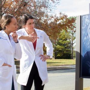 Jessica Waibl Polania (left) and Selena Lorrey, outside in their lab coats looking at a sign with the image of a brain on it. 