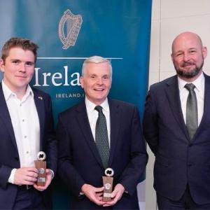 Donald McDonnell holding an award, standing between two other male recipients, in front of a sign that says Ireland with the Irish harp on it. 