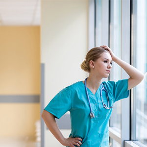 Woman in scrubs holding her head in exhaustion leaning against a window