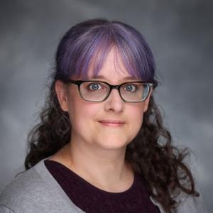 Headshot of a woman with a grey sweater, a purple shirt, black glasses and brown/purple hair.