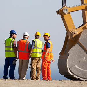 Four male construction workers in safety garb standing next to a giant backhoe shovel. 