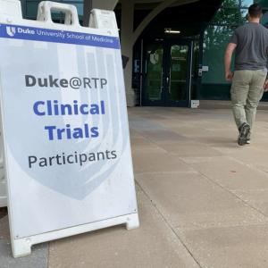placard on a sidewalk "Duke@RTP Clinical Trials Participants. Man in the background entering a building.