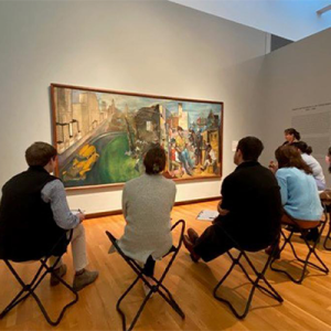 Residents sitting on folded stools in an art gallery looking at a large painting