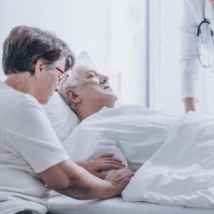 Elderly woman at the bedside of an elderly man in a hospital bed being treated by a healthcare worker.