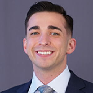 Headshot of Chris Stignham smiling at camera in front of grey backdrop wearing suit with silver tie