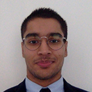 Headshot of Danyal Raza with close mouthed smile at camera, in front of white wall, wearing thin-rimmed glasses, blue shirt, black tie and black suit